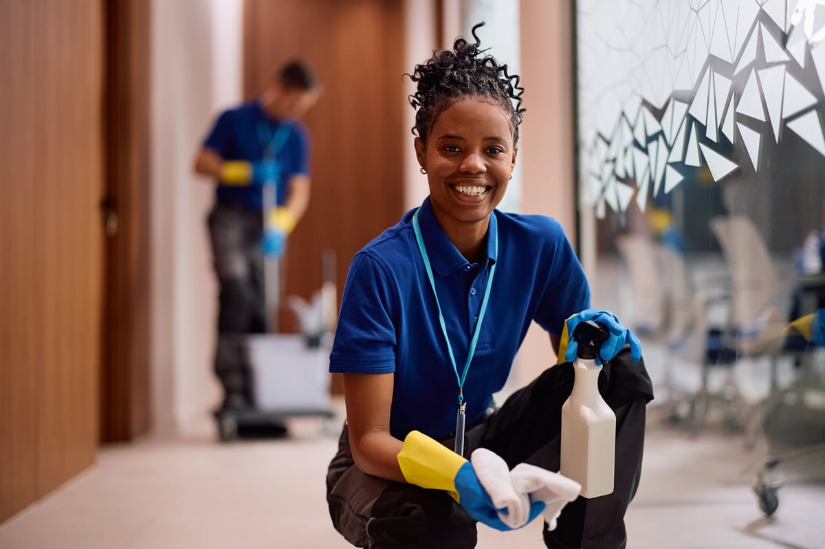 Happy African American female cleaner wiping glass walls in hallway of an office building and looking at camera